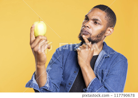 Young adult looks at an apple and thinks what can he cook with it, admiring colorful ripe fruit from local farmers market. Pensive man preparing to do a meal or dessert in studio. 117790678