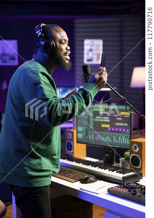 Talented performer singer does a recording session for his acoustic song, performing on microphone in home studio. Confident artist musician working on producing new track records. Talented performer singer does a recording session for his acoustic song, performing on microphone in home studio. Confident artist musician working on producing new track records. 117790716