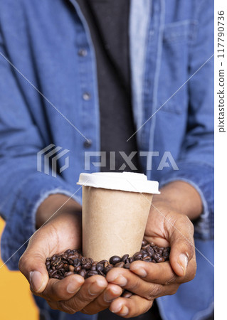 Young adult presenting cup of caffeine hot drink in front of the camera, brewing black roasted coffee beans. African american person serving cold brew beverage with delicious arabica flavor. Close up. Young adult presenting cup of caffeine hot drink in front of the camera, brewing black roasted coffee beans. African american person serving cold brew beverage with delicious arabica flavor. Close up. 117790736