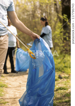 Activist using tongs tool to collect garbage and plastic waste from the forest, storing rubbish in a trash bag and clean the natural environment. Male volunteer grabbing litter and junk. 117790743