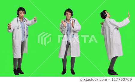 Happy woman specialist listening to cool music on headphones, having fun with modern tunes and dancing against greenscreen backdrop. Confident medic enjoys songs and shows dance moves. Camera A. 117790769