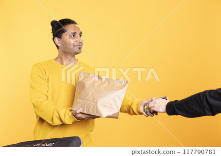 Jolly delivery worker offering groceries order to man paying with debit card. Customer buying meal from upbeat courier, swiping card on POS device, isolated over studio backdrop 117790781