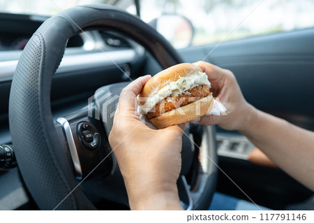 Asian lady holding hamburger to eat in car, dangerous and risk an accident. Asian lady holding hamburger to eat in car, dangerous and risk an accident. 117791146