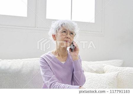Senior woman thinking while talking on smartphone in living room Senior woman thinking while talking on smartphone in living room 117791183