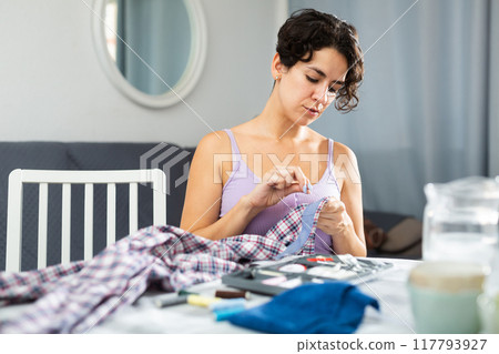 Portrait of attractive focused female using needle and thread to sew shirt 117793927