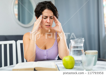 Young woman with headache sitting at table during reading book 117793958