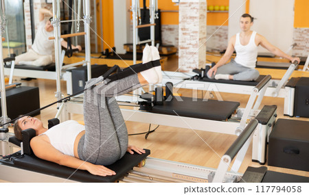 Girl works out in gym and performs set of exercises to stretch leg muscles with help of reformer 117794058