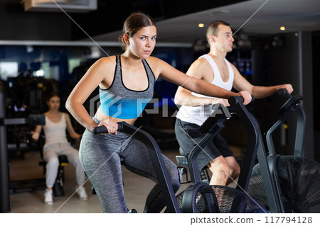 Sportive young couple doing air bike exercises in well-equipped gym Sportive young couple doing air bike exercises in well-equipped gym 117794128