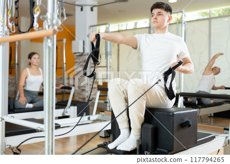 Man in gym with help of rope reformer performs exercises to strengthen muscles of arms 117794265