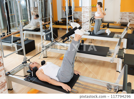 Man works out in gym and performs set of exercises to stretch leg muscles with help of reformer Man works out in gym and performs set of exercises to stretch leg muscles with help of reformer 117794296