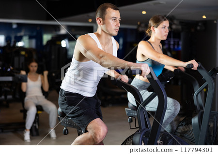 Guy performs exercises with stationary air bicycle in gym 117794301