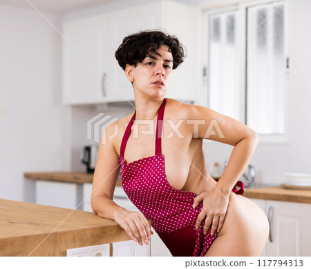 Sensual Latin woman in red apron dressed on naked corpus leaning on kitchen table at home 117794313