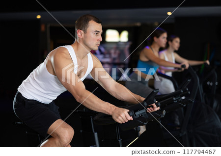 Sportive young guy doing spin bike exercises in well-equipped gym Sportive young guy doing spin bike exercises in well-equipped gym 117794467