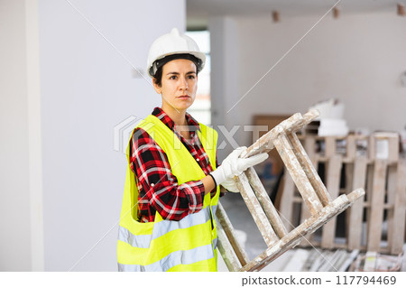 Young woman builder in construction helmet and vest carrying stepladder Young woman builder in construction helmet and vest carrying stepladder 117794469