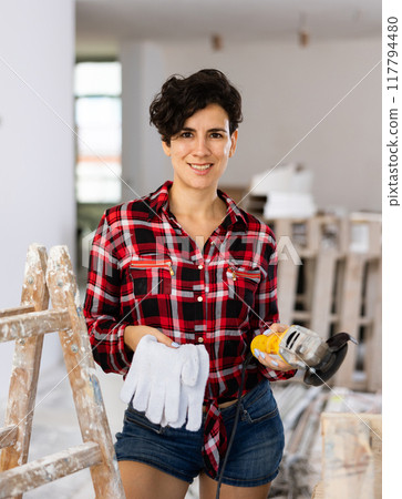 Pretty woman in a plaid shirt and short shorts poses playfully with a angle grinder in renovated room 117794480