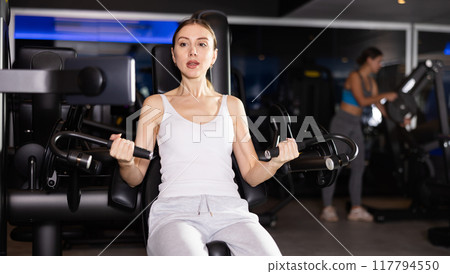 Sportive young girl training on arm curl machine in gym Sportive young girl training on arm curl machine in gym 117794550