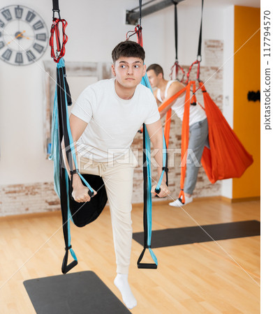 Man visitor performs exercises in hanging position on Pilates slings hammock 117794570