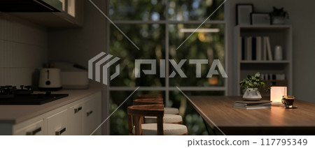 A side view image of a cozy, nighttime kitchen scene featuring a wooden dining table with stools. A side view image of a cozy, nighttime kitchen scene featuring a wooden dining table with stools. 117795349