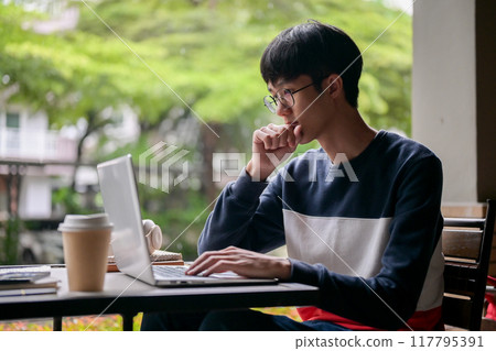 A thoughtful young Asian man is working remotely from an outdoor table, working on his laptop. 117795391