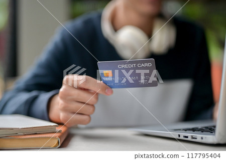 A close-up image of a man holding a credit card over a table while using his laptop. 117795404