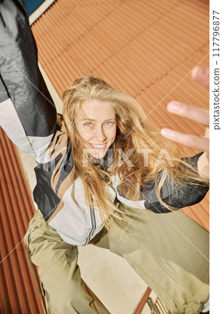Blonde woman with blue eyes smiling joyfully while climbing on metal structure against rust-colored backdrop 117796877