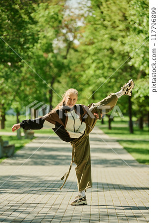 Person executing high kick balancing on one leg in serene urban park with green trees in background on sunny day wearing casual attire Person executing high kick balancing on one leg in serene urban park with green trees in background on sunny day wearing casual attire 117796899
