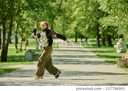 Young woman dancing energetically in the middle of park path surrounded by lush green trees and sunlight casting through leaves creating vibrant atmosphere Young woman dancing energetically in the middle of park path surrounded by lush green trees and sunlight casting through leaves creating vibrant atmosphere 117796901