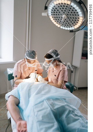 Vertical portrait of two trichologist specialist collecting hair follicles from donor area of male patient for hair transplantation in clinic. Process of hair transplant with FUE Sapphire method. 117799498