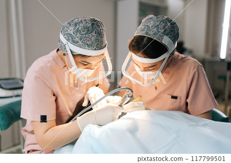 Closeup of two female doctors extracting hair follicles from donor area of patient for hair transplantation in clinic. Process of hair transplant with FUE method. Concept of baldness treatment. 117799501