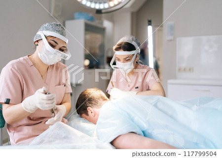 Two female trichologist performing local needle-free anesthesia to back of head using pistol in recipient area prior to hair transplant surgery in clinic. Concept of baldness treatment 117799504