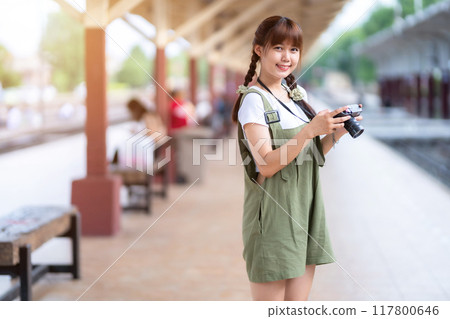 Portrait Young asian woman smiling tourist Traveler girl walking and with a holding the camera waits train travel journey is taken in railway platform Thailand, summer relax vacation Concept. 117800646
