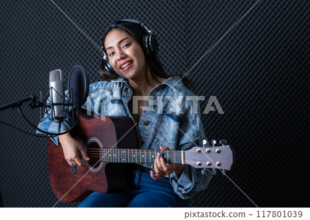 Happy cheerful pretty smiling of portrait a young Asian woman vocalist Wearing Headphones with a Guitar recording a song front of microphone in a professional studio 117801039