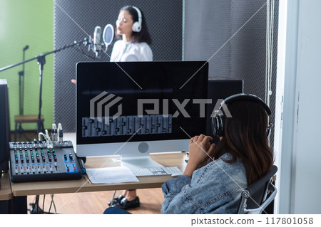 female studio technician of the computer monitor of Software for recording in foreground of young Asian woman vocalist Wearing Headphones recording a song front of microphone in a professional studio female studio technician of the computer monitor of Software for recording in foreground of young Asian woman vocalist Wearing Headphones recording a song front of microphone in a professional studio 117801058