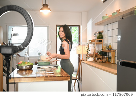 Athletic female preparing a nutritious recipe and recording video with phone camera at home kitchen 117807009