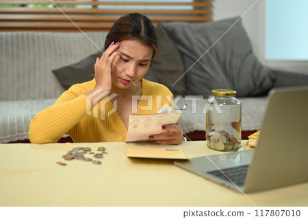 Stressed young woman managing expenses and planning personal budget while sitting in living room Stressed young woman managing expenses and planning personal budget while sitting in living room 117807010