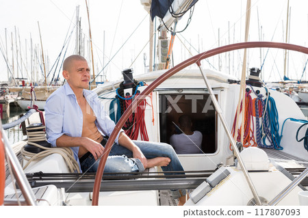 European guy with a naked torso and a bottle of beer in his hand sits on the deck of a yacht in the port 117807093