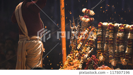 Varanasi, Uttar Pradesh, India. Man making movements with a flowers petals on the Ganga Maha Aarti ceremony begins. Brahman priest wearing red and yellow dress doing traditional ganga aarti at ghat 117807196