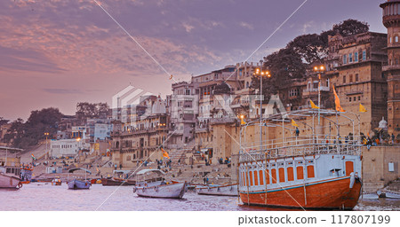 Varanasi, India. General View From Pleasure Boat on Chausathi Ghat. Purple Sunset Sky At Evening . Many Boats Moored On Ganga River. Devotees Take A Holy Dip In Water From This Ghat Varanasi, India. General View From Pleasure Boat on Chausathi Ghat. Purple Sunset Sky At Evening . Many Boats Moored On Ganga River. Devotees Take A Holy Dip In Water From This Ghat 117807199