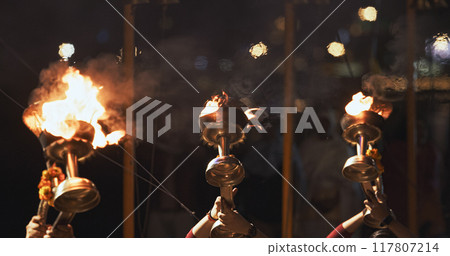Varanasi, India. Man makes movements with a candle holder with lit candles on the Ganga Maha Aarti ceremony begins. Brahman priest wearing red and yellow dress doing traditional ganga aarti at ghat Varanasi, India. Man makes movements with a candle holder with lit candles on the Ganga Maha Aarti ceremony begins. Brahman priest wearing red and yellow dress doing traditional ganga aarti at ghat 117807214