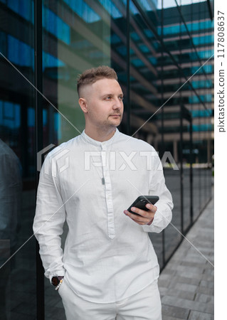 Focused young corporate worker investor businessman walking and using mobile phone outdoors near a business centre.  117808637