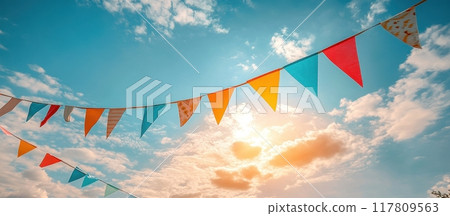 Colorful Festive Bunting Flags Against a Bright Blue Sky with Clouds and Sunlight 117809563