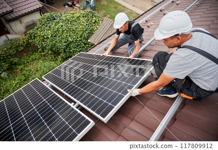 Men technicians mounting photovoltaic solar moduls on roof of house. Engineers in helmets installing solar panel system outdoors. Concept of alternative and renewable energy. Men technicians mounting photovoltaic solar moduls on roof of house. Engineers in helmets installing solar panel system outdoors. Concept of alternative and renewable energy. 117809912
