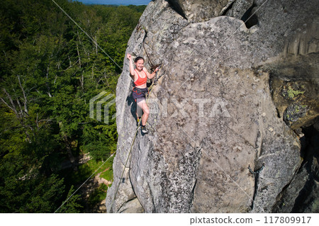 Aerial view of woman rock climber ascending rugged limestone cliff with harness and rope for safety. Sportswoman climbing on vertical large boulder at Dobvush Rocks in Carpathian mountains, Ukraine. Aerial view of woman rock climber ascending rugged limestone cliff with harness and rope for safety. Sportswoman climbing on vertical large boulder at Dobvush Rocks in Carpathian mountains, Ukraine. 117809917