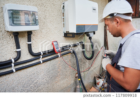 Man electrician installing solar panel system. Technician in helmet and gloves making electrical wiring inverter and electric box. Concept of alternative and renewable energy. 117809918