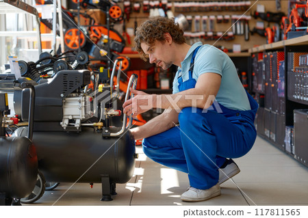 Young workman in overalls choosing pneumatic compressor at hardware store Young workman in overalls choosing pneumatic compressor at hardware store 117811565