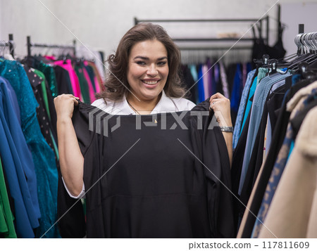 A fat woman in a plus size store tries on a black evening dress. Vertical photo.  117811609