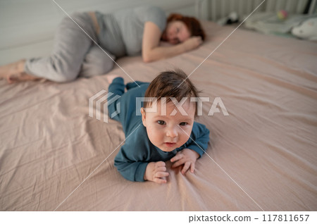A three-month-old boy lies on his stomach on the bed and his mother sleeps behind him. Postpartum depression. 117811657