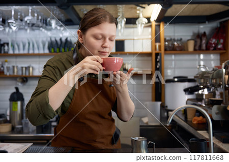 Satisfied woman barista smelling freshly brewed coffee in ceramic cup 117811668