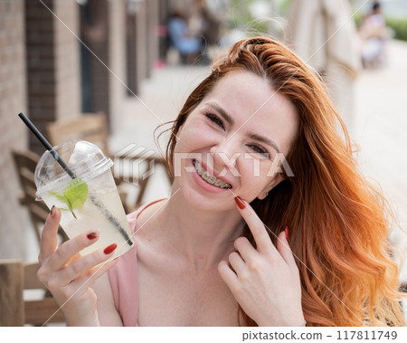 Young beautiful red-haired woman with braces drinks cooling lemonade outdoors in summer. Portrait of a smiling girl with freckles. 117811749