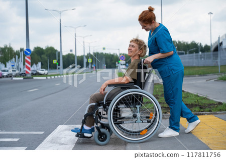 Red-haired nurse pushing an elderly woman in a wheelchair across the road. Red-haired nurse pushing an elderly woman in a wheelchair across the road. 117811756